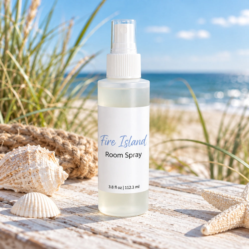 A Fire Island room spray stands on a weathered wooden surface outdoors with seashells, rope, beach grass, and the ocean in the background. The frosted plastic bottle has a white spray top and a white label featuring blue and black text.