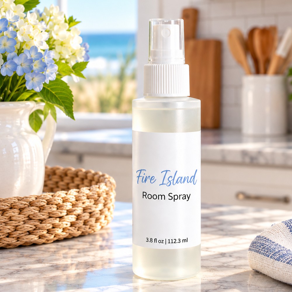 A frosted plastic room spray bottle labeled 'Fire Island Room Spray' stands on a marble countertop in a bright kitchen setting with flowers, a basket, and a blurred window view in the background.