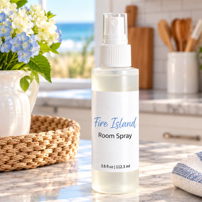 A frosted plastic room spray bottle labeled 'Fire Island Room Spray' stands on a marble countertop in a bright kitchen setting with flowers, a basket, and a blurred window view in the background.