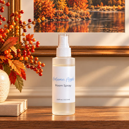 A bottle of Autumn Apple room spray stands on a wooden table in a warm, autumn-themed setting. The scene includes a vase of orange berries on the left and a framed picture of an autumn forest in the background.