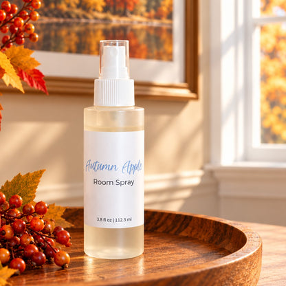 A bottle of Autumn Apple Room Spray stands on a wooden tray in a warm, autumn themed setting with fall leaves and berries, and a window in the background.