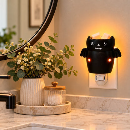 Halloween bat candle wax warmer plugged into a bathroom wall with plant  and glass container besides it.