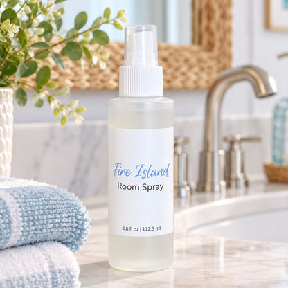 A frosted room spray plastic bottle labeled 'Fire Island Room Spray' stands on a bathroom counter near a sink, with folded towels, a plant, and a woven mirror in the background.