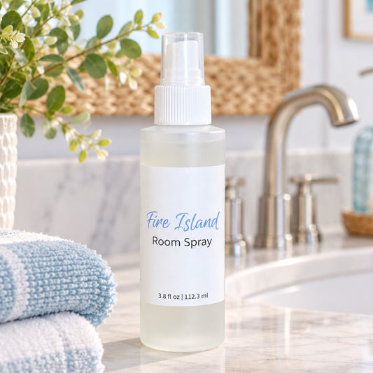 A frosted room spray plastic bottle labeled 'Fire Island Room Spray' stands on a bathroom counter near a sink, with folded towels, a plant, and a woven mirror in the background.