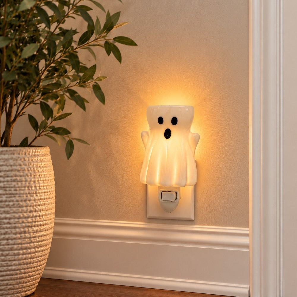 A ghost shaped wax warmer plugged into a wall outlet, glowing with a warm yellow light. The ghost has a white body with black eyes and mouth.