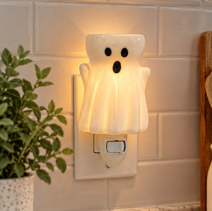 A ghost shaped plug in white candle wax melt warmer plugged into a outlet against a beige subway tile.  There is a green plant on the left and a wooden object on the right.