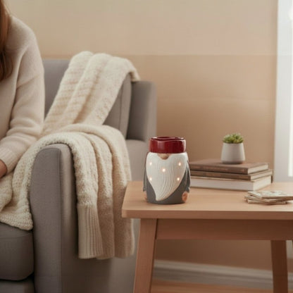 Person sitting on a couch with a blanket, next to a wooden table with a gnome candle wax fragrance warmer.