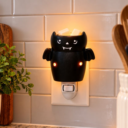 A black plug-in wax melt warmer with glowing orange eyes and a warm yellow light, shown plugged into a white outlet against a beige tiled wall with kitchen props nearby.
