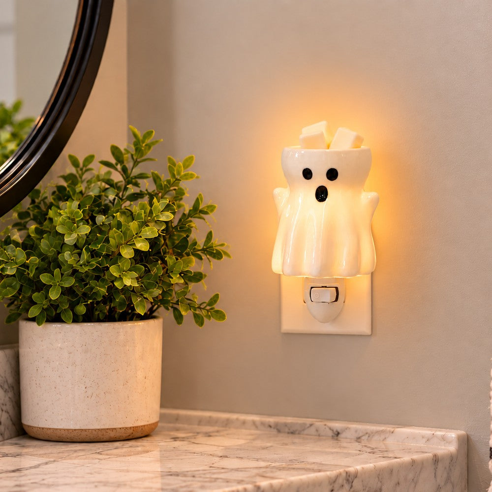 A ghost shaped wax warmer plugged into a wall outlet on a beige wall, glowing warm white with black facial features. A potted green plant and part of a mirror are visible on the left, and a marble countertop is below.