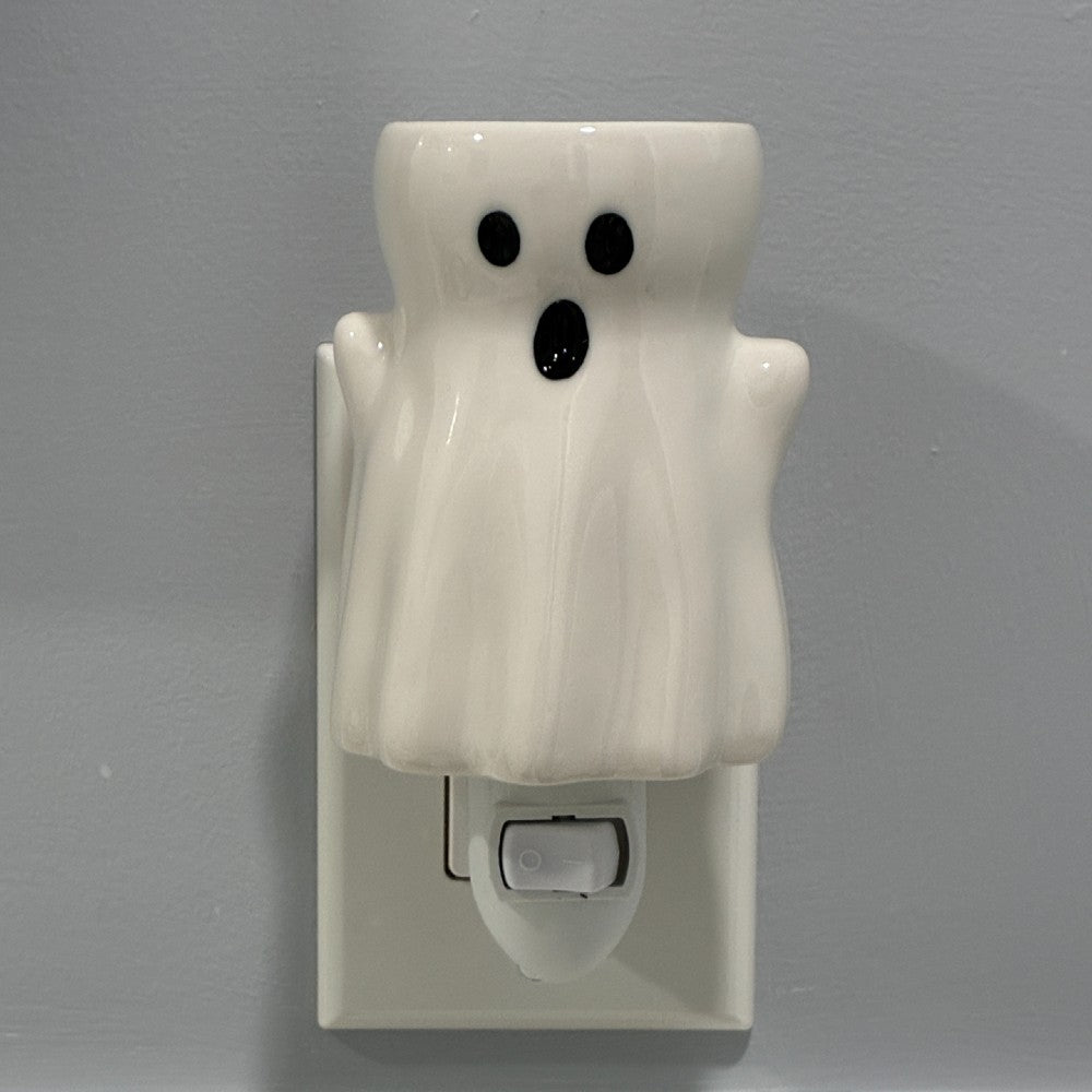 A white ceramic ghost shaped plug in candle wax melt warmer with black eyes and mouth.  It's shown plugged into a white wall outlet against a gray wall.