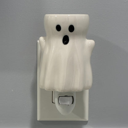 A white ceramic ghost shaped plug in candle wax melt warmer with black eyes and mouth.  It's shown plugged into a white wall outlet against a gray wall.