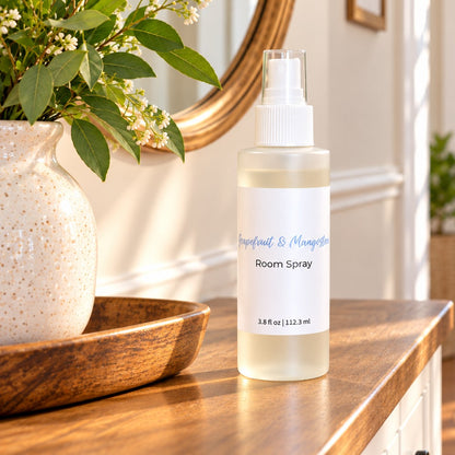 A frosted plastic room spray bottle with a white spray top and a white label reading Grapefruit & Mangosteen room spray sits on a wooden table in a bright, styled interior. A speckled ceramic vase with green leaves and small white flowers is on a wooden tray beside it.
