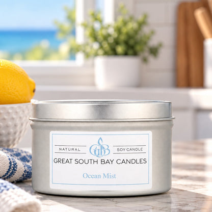 A silver metal tin candle labeled 'Great South Bay Candles Ocean Mist' sits on a countertop in a bright kitchen setting with a lemon, plant, and window in the background.