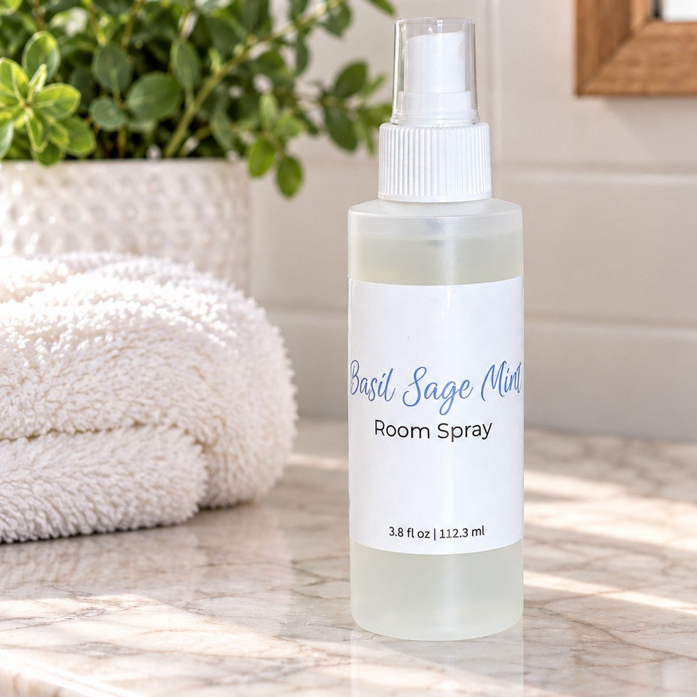 A frosted room spray bottle labeled 'Basil Sage Mint Room Spray' standing on a marble surface with a folded white towel and green plant in the background.