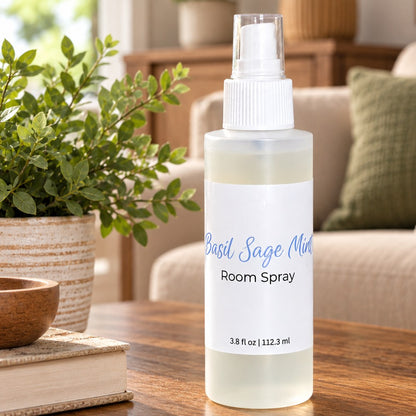 A bottle of Basil Sage Mist Room Spray stands on a wooden table in a cozy living room setting, with a potted plant and books in the foreground and a sofa in the background.