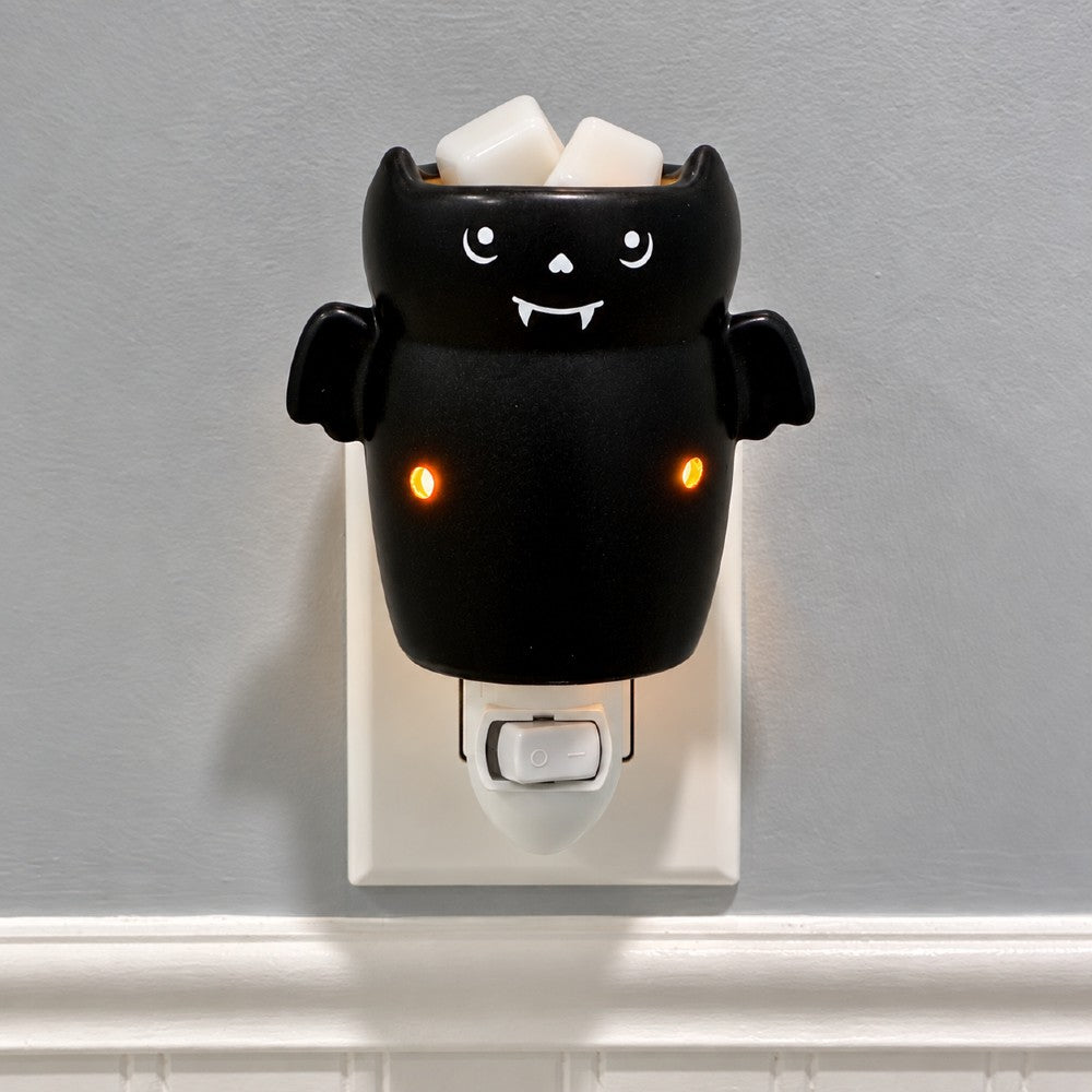 A black bat shaped plug in candle wax warmer with glowing orange eyes. Shown mounted on a wall outlet against a light gray wall. 
