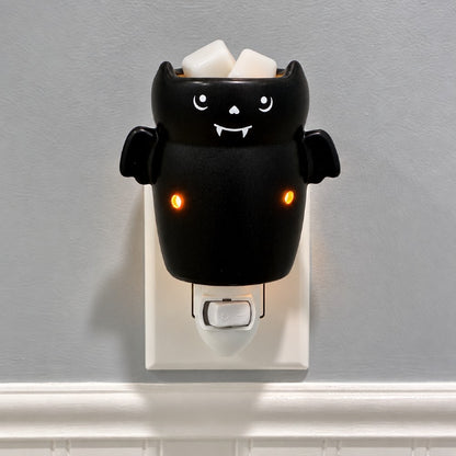 A black bat shaped plug in candle wax warmer with glowing orange eyes. Shown mounted on a wall outlet against a light gray wall. 