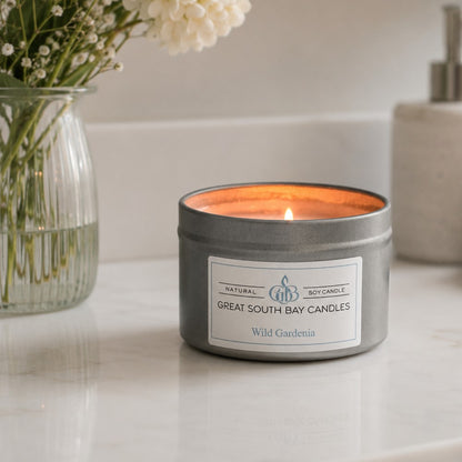 A lit silver metal tin candle labeled 'Great South Bay Candles' and 'Wild Gardenia' sits on a white marble countertop. A glass vase with white flowers is blurred in the background on the left, and a neutral bathroom or home object is visible on the right.