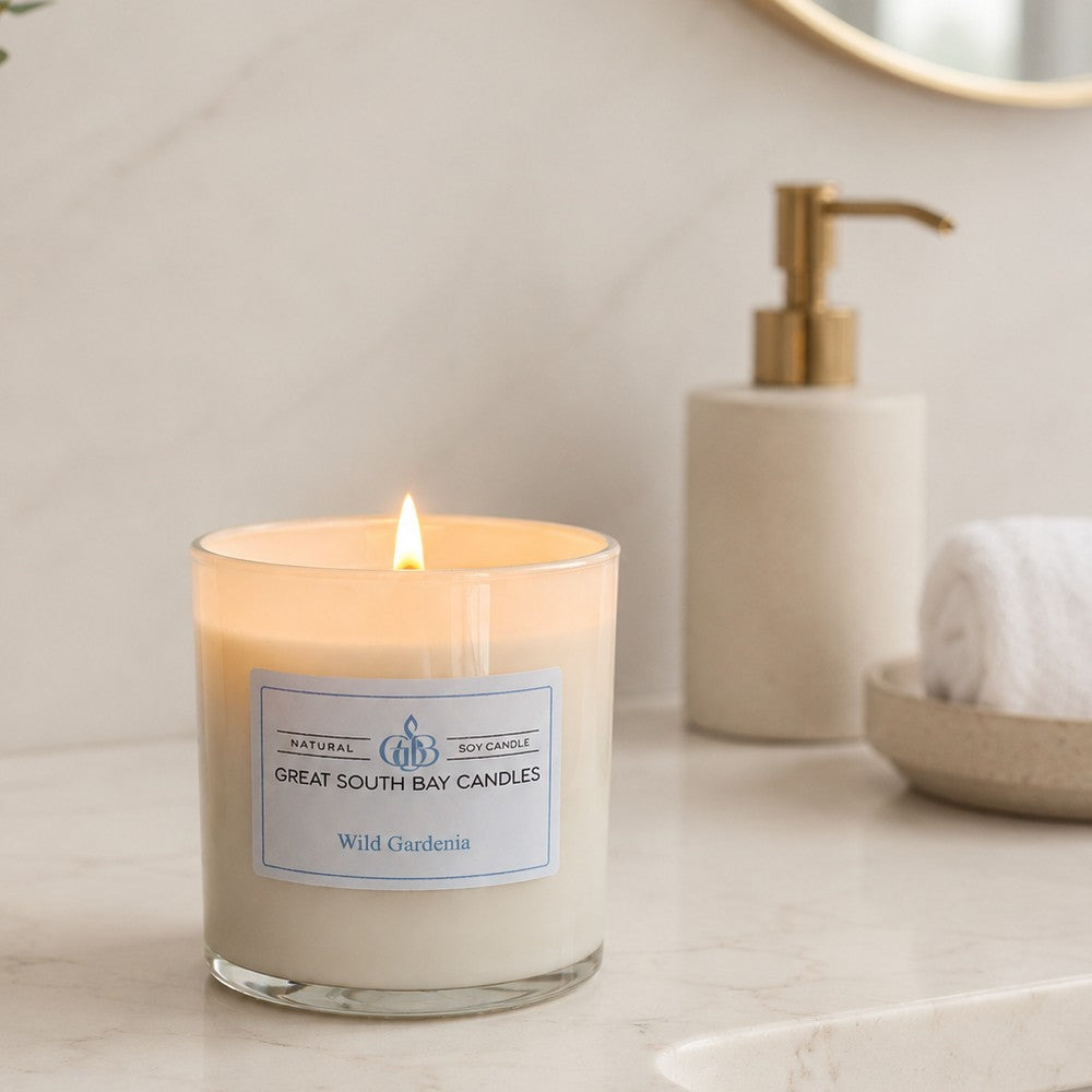 A lit soy candle in a white glass jar sits on a light countertop beside a soap dispenser. The candle label reads 'Natural Soy Candles' and 'Wild Gardenia'.